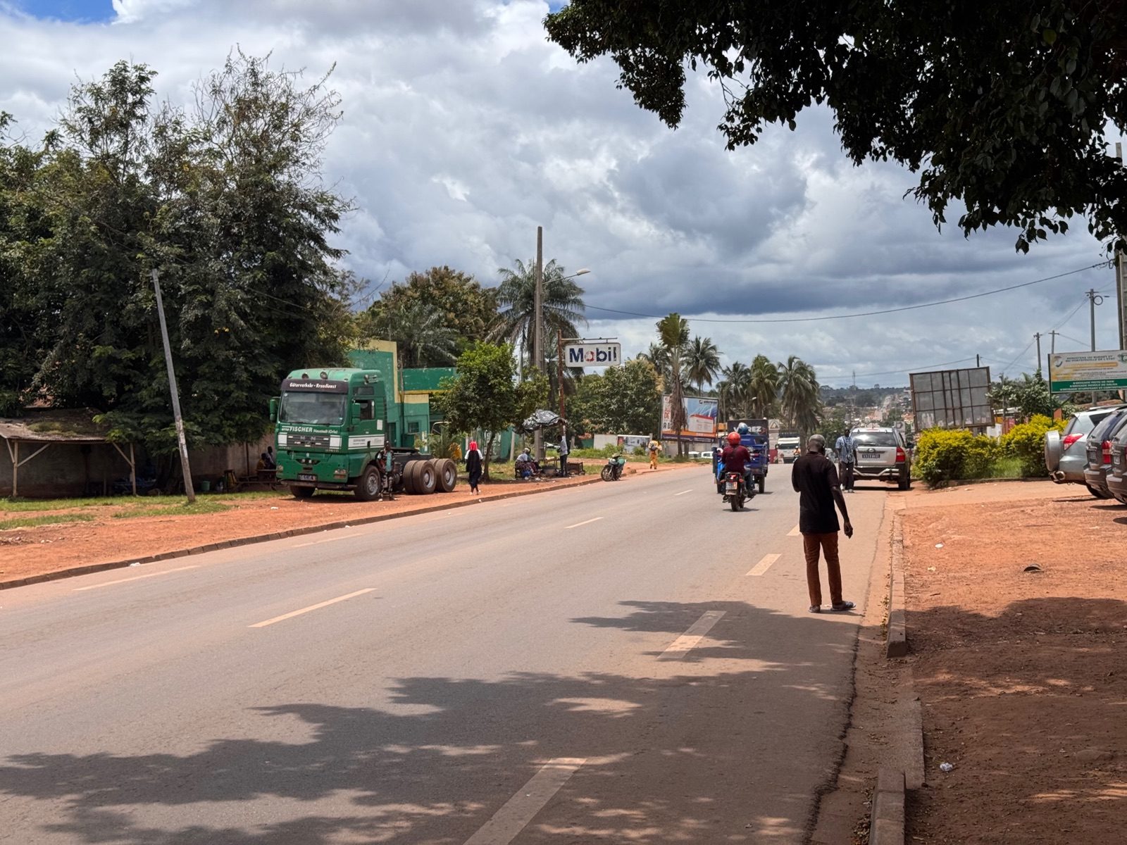 Main street of an inner Côte d'Ivoire town: truck parked by a fuel station, motorbikes and pedestrians under a rainy-season sky