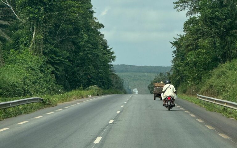 Paved highway through tropical forest in Côte d'Ivoire, a motorbike and a loaded cart heading towards plantations on the horizon