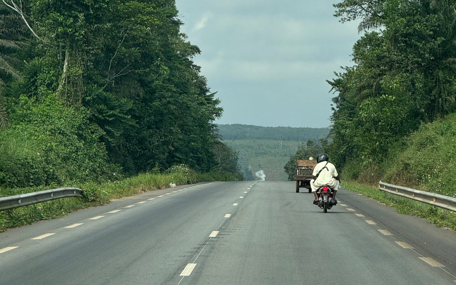 Paved highway through tropical forest in Côte d'Ivoire, a motorbike and a loaded cart heading towards plantations on the horizon